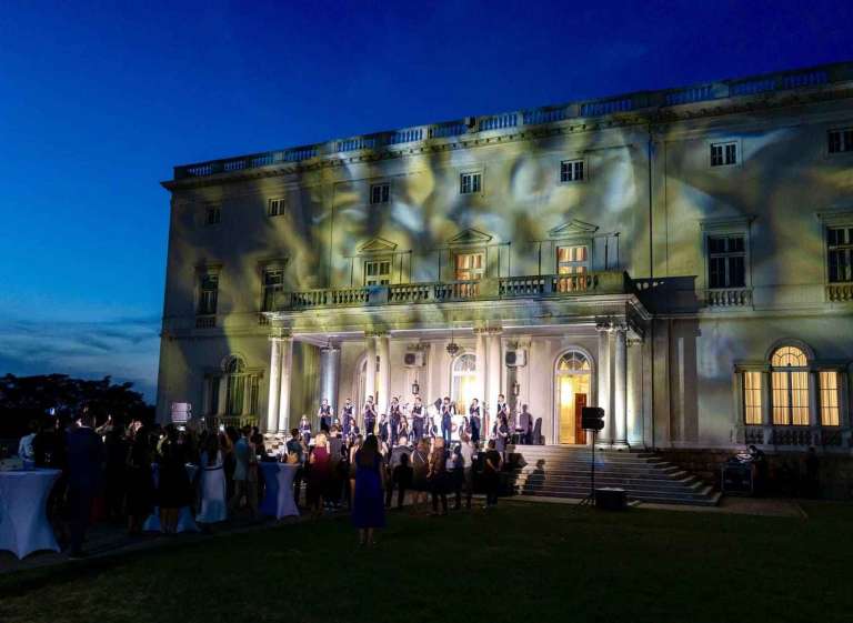 Wide shot of guests gathered on the lawn watching a performance at the White Palace during the IMGL 2026 event
