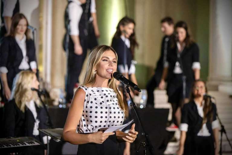 A speaker holding a microphone and notes on stage with the choir performing in the background at the White Palace gala